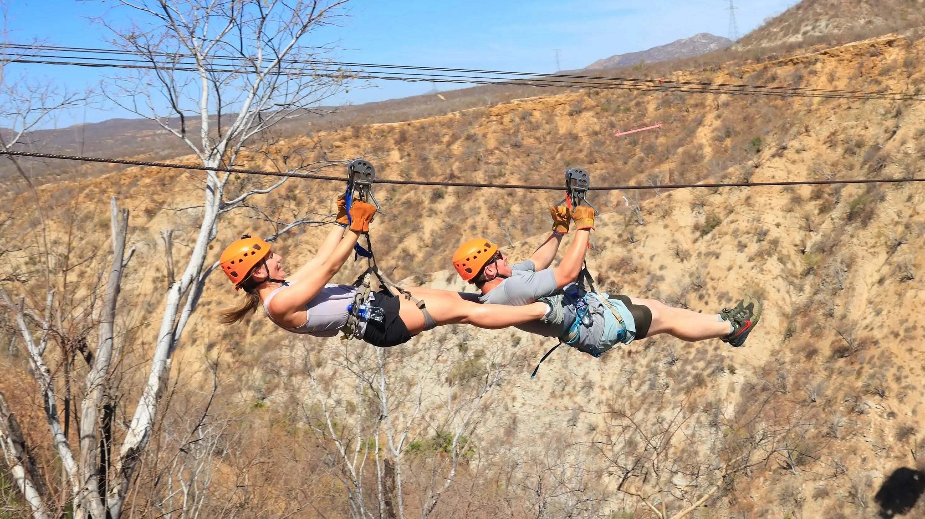Two guests in orange helmets riding a tandem zipline across the canyon at Wild Canyon in Los Cabos
