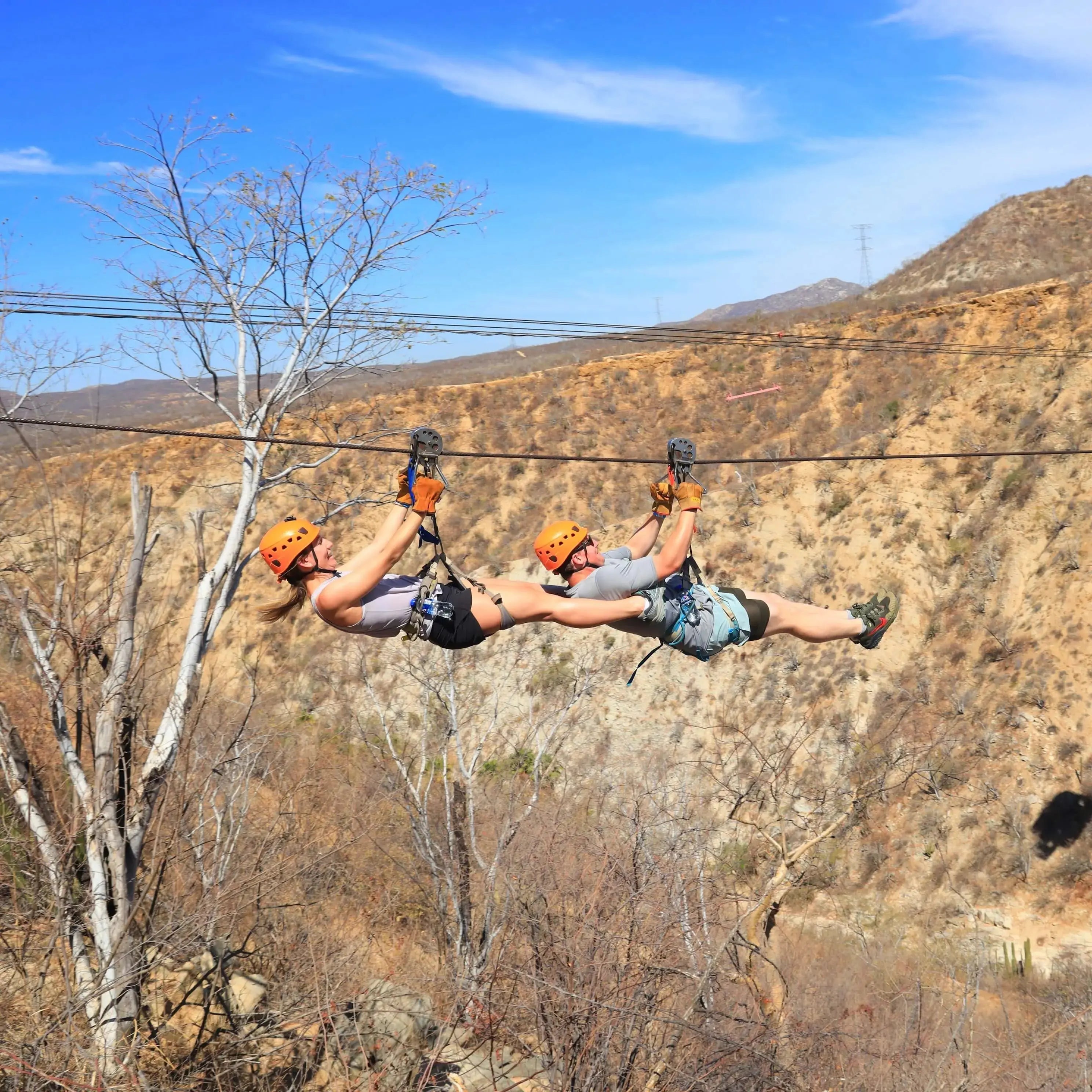 Two guests in orange helmets riding a tandem zipline across the canyon at Wild Canyon in Los Cabos